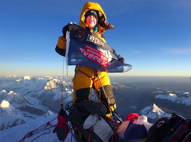 Melanie Windridge in climbing gear at the summit of Mount Everest, holding a 'science of Everest' banner