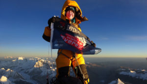 Melanie Windridge in climbing gear at the summit of Mount Everest, holding a "science of Everest" banner